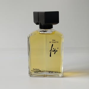 May include: A clear glass perfume bottle filled with golden liquid, featuring a black rectangular cap. The front label displays the text Eau de Toilette, Fidji, Parfums Guy Laroche Paris, and 57 ml - 85% vol. The bottle has a distinct angular shape.