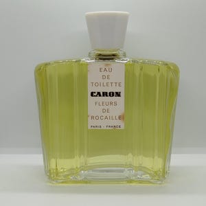 Può includere: Un flacone di profumo rettangolare in vetro riempito con liquido giallo pallido, dotato di un tappo bianco e un'etichetta centrale che riporta EAU DE TOILETTE CARON FLEURS DE ROCAILLE PARIS FRANCE. Il flacone presenta scanalature verticali.