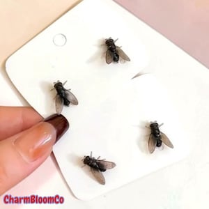 May include: Four small, realistic fly-shaped stud earrings with black bodies and translucent, smoky-grey wings. Each fly is mounted on a white rectangular display card, featuring detailed insect anatomy including visible legs and segmented bodies.
