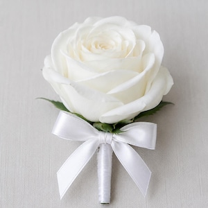 Könnte beinhalten: Ein einzelnes Ansteckblümchen mit einer weißen Rose in voller Blüte und grünen Kelchblättern an der Basis. Der Stiel ist mit weißem Satinband umwickelt und mit einer passenden Schleife aus weißem Satinband versehen, die ordentlich gebunden ist.