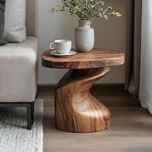 Puede incluir: Una mesa auxiliar de madera con una superficie lisa y redondeada y una base escultural retorcida con vetas de madera visibles. Una taza y un plato de cerámica blanca se encuentran sobre la mesa junto a un jarrón de cerámica gris claro.