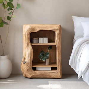 May include: A light brown wooden nightstand with a natural, irregular live-edge frame. The piece features two open shelves containing a small grey bowl, a stack of books, and a potted green plant with large leaves, showcasing a raw wood texture.