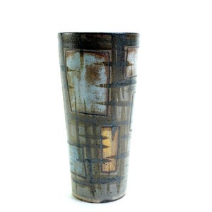 May include: A cylindrical ceramic vase featuring a dark brown base with a textured, grid-like pattern of intersecting lines. The surface is decorated with rectangular patches of muted blue and tan glazes, creating a layered, geometric appearance.
