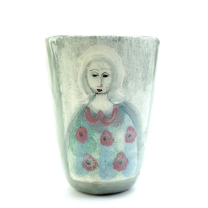 May include: A ceramic tumbler with a tapered shape and a glossy finish. The exterior features a hand-painted illustration of a figure with light hair, wearing a blue garment adorned with a repeating pattern of small red flowers and a pink collar.