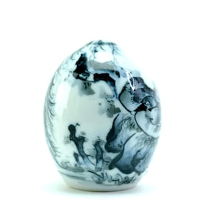 Può includere: Un piccolo vaso in ceramica a forma di uovo con una superficie bianca liscia e lucida. Presenta illustrazioni dipinte a mano con inchiostro blu scuro di elementi paesaggistici astratti, tra cui alberi stilizzati e formazioni rocciose.