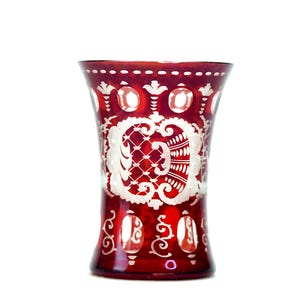 Könnte beinhalten: Eine rubinrote Glasvase mit weißen geätzten floralen und geometrischen Mustern. Die Vase hat einen ausgestellten oberen Rand und einen bauchigen Boden. Das weiße Design umfasst ovale Formen und ein zentrales verziertes Muster. Die Vase ist wahrscheinlich Vintage oder antik.