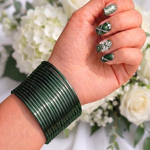 May include: A stack of thin, metallic dark green bangles worn on the wrist. The bangles have a smooth, reflective finish and a uniform circular shape, creating a wide, layered band of deep green color that encircles the lower arm.