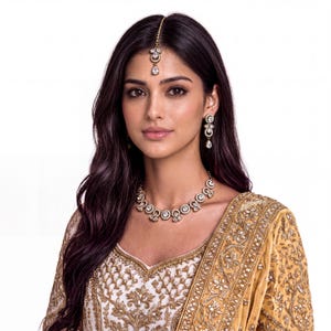 May include: A gold-toned jewelry set featuring a necklace, drop earrings, and a maang tikka headpiece. Each piece is adorned with clear, pear-shaped and round crystals set in a metallic frame, paired with a gold-embroidered white and tan garment.