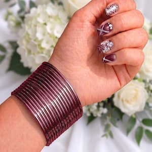 May include: A set of numerous thin, metallic maroon bangles worn together on the wrist. The bracelets feature a smooth, reflective finish and a uniform cylindrical shape, creating a wide, stacked appearance that covers a portion of the forearm.