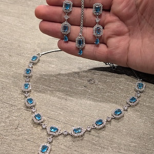 May include: A silver-toned jewellery set featuring a necklace, two drop earrings, and a maang tikka. Each piece is adorned with rectangular blue stones surrounded by small clear crystals, with a small teardrop-shaped blue stone at the base.