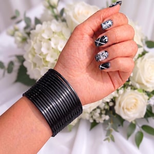 May include: A set of numerous thin, glossy black bangles worn together on the wrist. The bracelets are uniform in color and texture, creating a wide, stacked appearance that covers a portion of the forearm with a smooth, reflective finish.