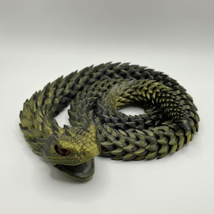 May include: A coiled articulated snake figurine featuring a textured, scale-like surface in a gradient of metallic olive green and matte black. The snake has a wide, open mouth and prominent, dark red eyes, with a segmented body design.