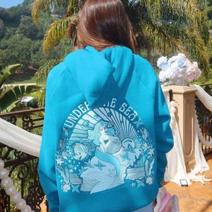 May include: A turquoise hoodie featuring a graphic of a mermaid, fish, and the words "UNDER THE SEA." The design is set within a circular frame with a sunburst effect. The hoodie is worn by a person in blue jeans.