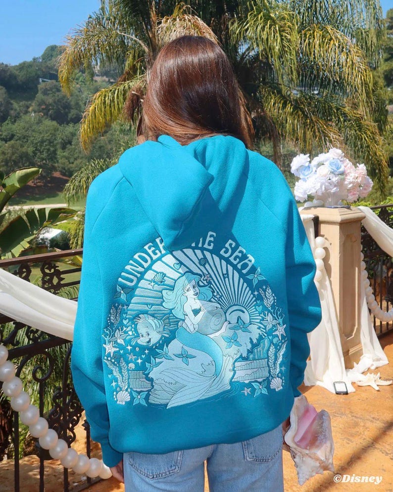 May include: A turquoise hoodie featuring a graphic of a mermaid, fish, and the words "UNDER THE SEA." The design is set within a circular frame with a sunburst effect. The hoodie is worn by a person in blue jeans.