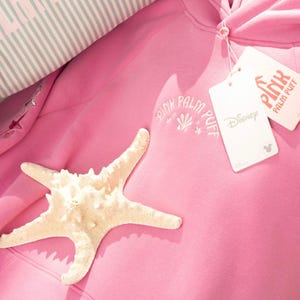 May include: A light pink hoodie with the text "PINK PALM PUFF" and a seashell graphic. A white starfish rests on the hoodie. A tag with the Disney logo is attached. A striped pillow is visible in the background.