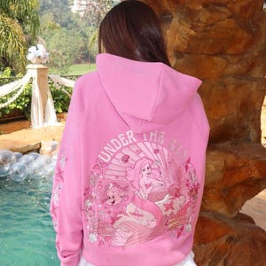 May include: Pink hooded sweatshirt with a graphic of a mermaid and the text "UNDER THE SEA". The design includes shells, flowers, and other sea creatures. The sleeves have additional floral accents.