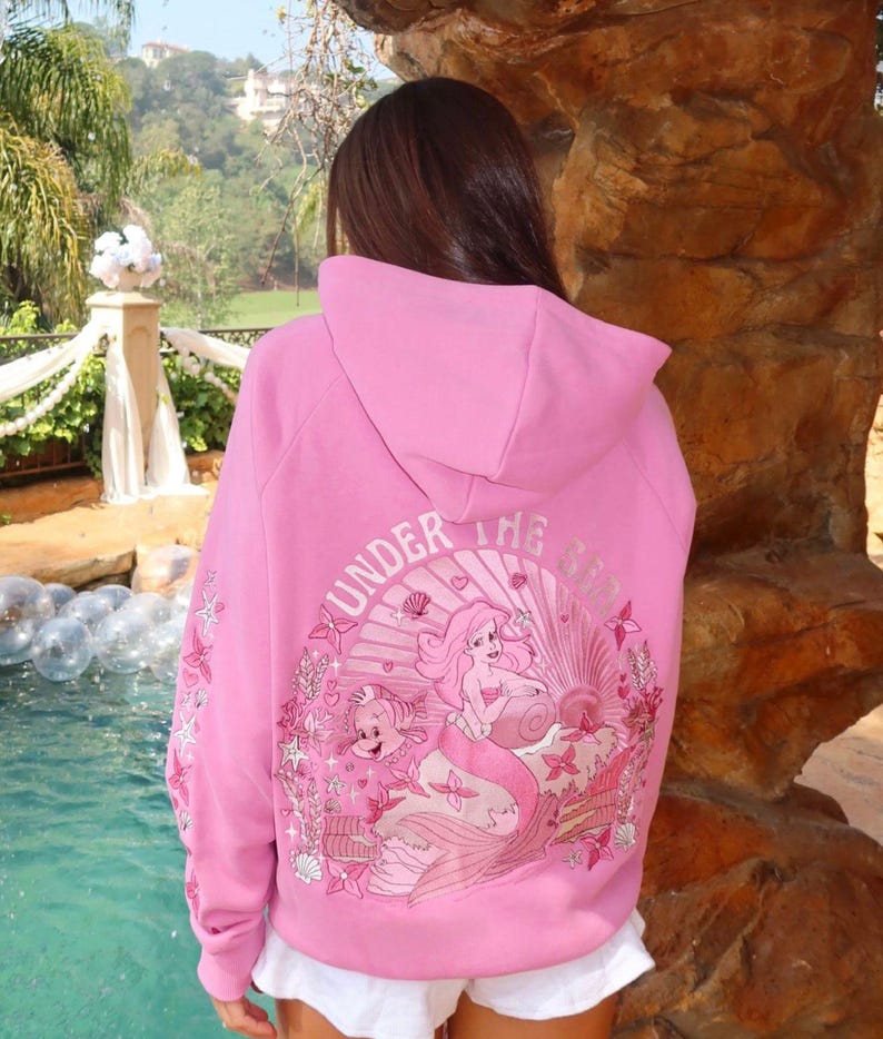 May include: Pink hooded sweatshirt with a graphic of a mermaid and the text "UNDER THE SEA". The design includes shells, flowers, and other sea creatures. The sleeves have additional floral accents.