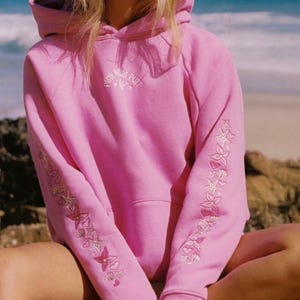 May include: A pink hooded sweatshirt with embroidered floral and seashell designs on the sleeves. The text "Pink Palm Puff" is embroidered on the chest. The sweatshirt is made of a soft material and has a drawstring hood.