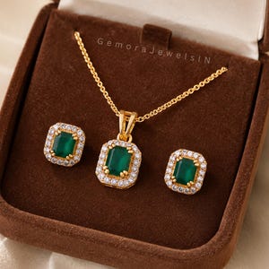 Puede incluir: Un conjunto de joyería en tono dorado que incluye un collar y pendientes a juego, cada uno con una piedra rectangular verde rodeada de cristales blancos. El conjunto se presenta en una caja marrón con el texto GemoraJewelsIN impreso.