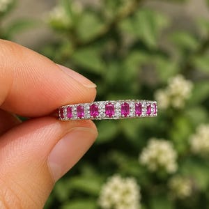 May include: A silver-toned band ring featuring a row of alternating rectangular-cut vibrant pink stones and small clear round stones. The stones are set in a continuous line across the front of the metal band, creating a sparkling, multi-faceted appearance.