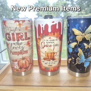 May include: Three stainless steel tumblers with lids. The left tumbler has a wood grain background, autumn leaves, a pumpkin, and the text "Just a girl who loves pumpkins." The centre tumbler features a pumpkin spice latte graphic and text. The right tumbler displays blue and yellow butterflies on a dark background. The text "New Premium Items" appears at the top.