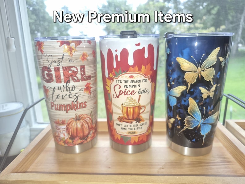 May include: Three stainless steel tumblers with lids. The left tumbler has a wood grain background, autumn leaves, a pumpkin, and the text "Just a girl who loves pumpkins." The centre tumbler features a pumpkin spice latte graphic and text. The right tumbler displays blue and yellow butterflies on a dark background. The text "New Premium Items" appears at the top.