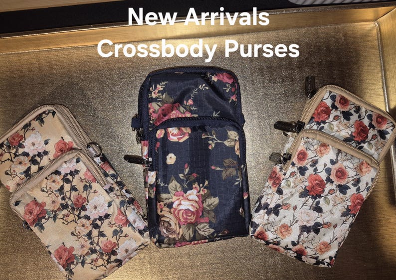 May include: Three rectangular crossbody purses featuring floral patterns. The left purse is beige, the centre is navy blue, and the right is white. Each purse has multiple zippered compartments and metal hardware. Text at the top reads: New Arrivals Crossbody Purses.