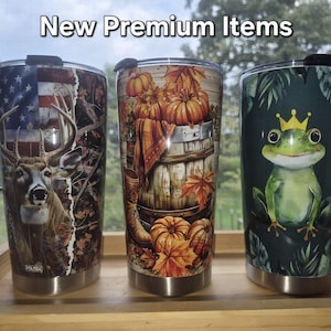 May include: Three stainless steel tumblers with lids, featuring the text New Premium Items. The left tumbler shows a deer and American flag, the centre features pumpkins and autumn leaves on a barrel, and the right displays a green frog with a crown.