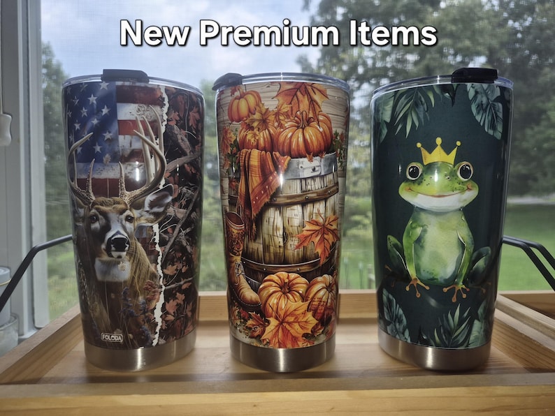 May include: Three stainless steel tumblers with lids, featuring the text New Premium Items. The left tumbler shows a deer and American flag, the centre features pumpkins and autumn leaves on a barrel, and the right displays a green frog with a crown.