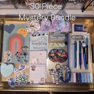 May include: A 30 Piece Mystery Bundle set arranged on a gold tray, including a rainbow card, Hyaluronic Acid mask, hair accessories, stickers, pens, a notepad, a craft ink pad, a keychain, and various stationery items in blue and purple tones.