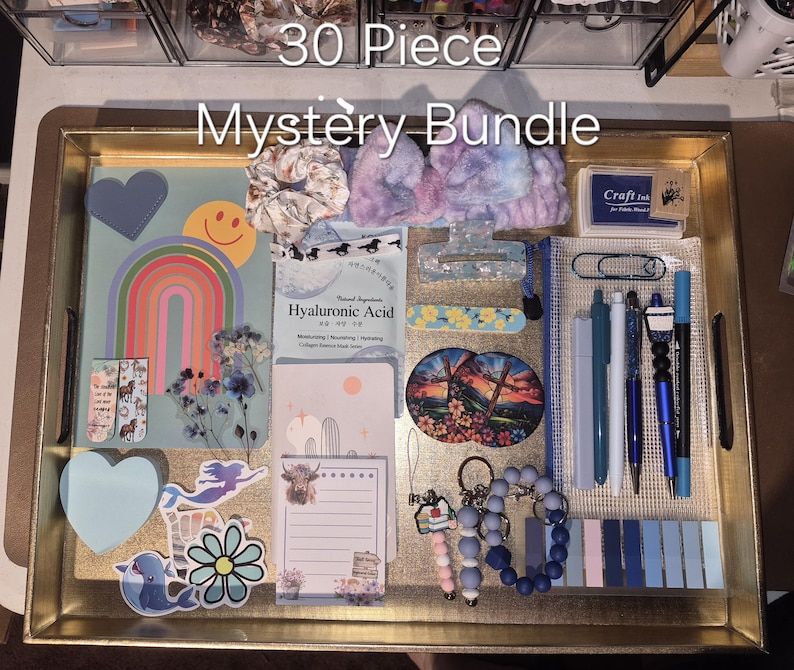 May include: A 30 Piece Mystery Bundle set arranged on a gold tray, including a rainbow card, Hyaluronic Acid mask, hair accessories, stickers, pens, a notepad, a craft ink pad, a keychain, and various stationery items in blue and purple tones.
