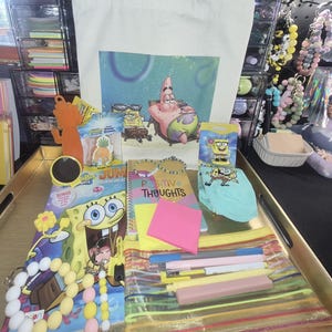 May include: A collection of SpongeBob SquarePants themed items including a canvas tote bag, a colouring book, a spiral notebook labelled Positive Thoughts, a pair of light blue socks, a clear pencil case with pens, and various silicone bead keychains.