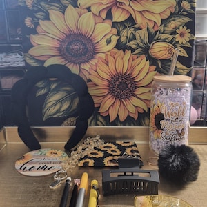 May include: A collection of items including a black tote bag with a large yellow sunflower print, a black padded headband, a glass tumbler with a bamboo lid and sunflower graphic, a round coaster, a black hair claw, pens, and a black pom-pom.