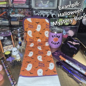 May include: A Halloween mystery bundle featuring an orange towel with white ghosts and the text 'hey boo!', a purple tumbler with a pumpkin and 'trick treat' text, a glass jar, pencils, a brain and eyeball toy, a spider, and various stationery items.