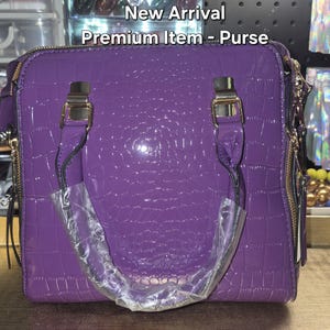 May include: A purple handbag with a glossy crocodile-embossed texture, featuring gold-tone metal hardware, two top handles, and side zips. The text New Arrival Premium Item - Purse is visible at the top of the image, with one handle wrapped in plastic.