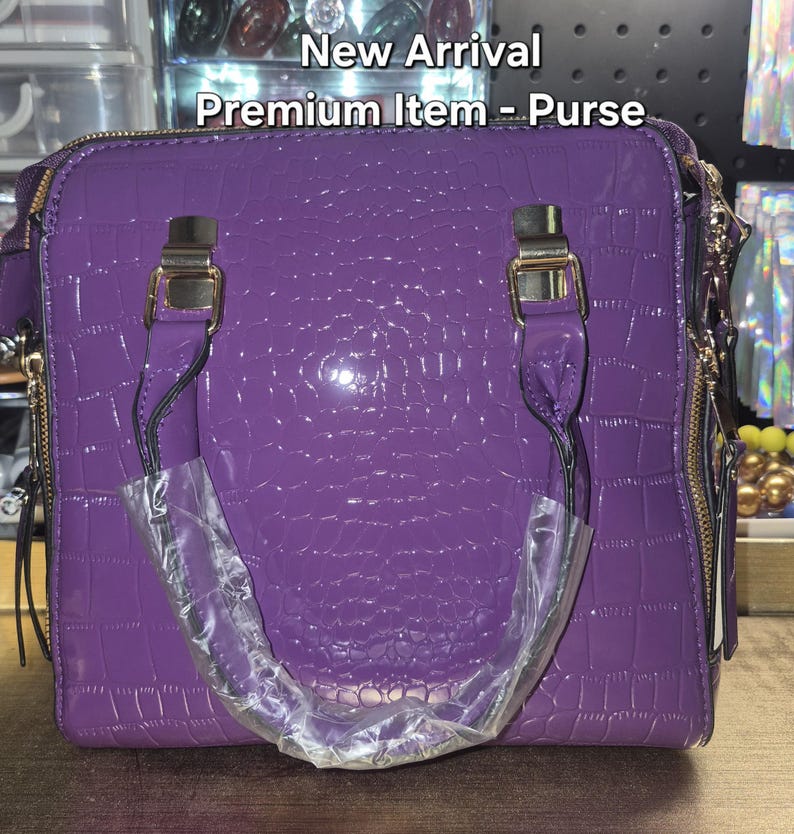 May include: A purple handbag with a glossy crocodile-embossed texture, featuring gold-tone metal hardware, two top handles, and side zips. The text New Arrival Premium Item - Purse is visible at the top of the image, with one handle wrapped in plastic.