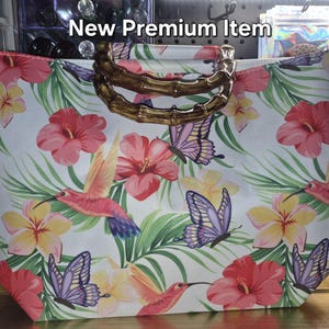 May include: A white tote bag featuring a vibrant tropical print of pink hibiscus flowers, yellow plumeria, green palm leaves, hummingbirds, and purple butterflies. The bag includes two curved, brown bamboo-style handles. Text reads New Premium Item.