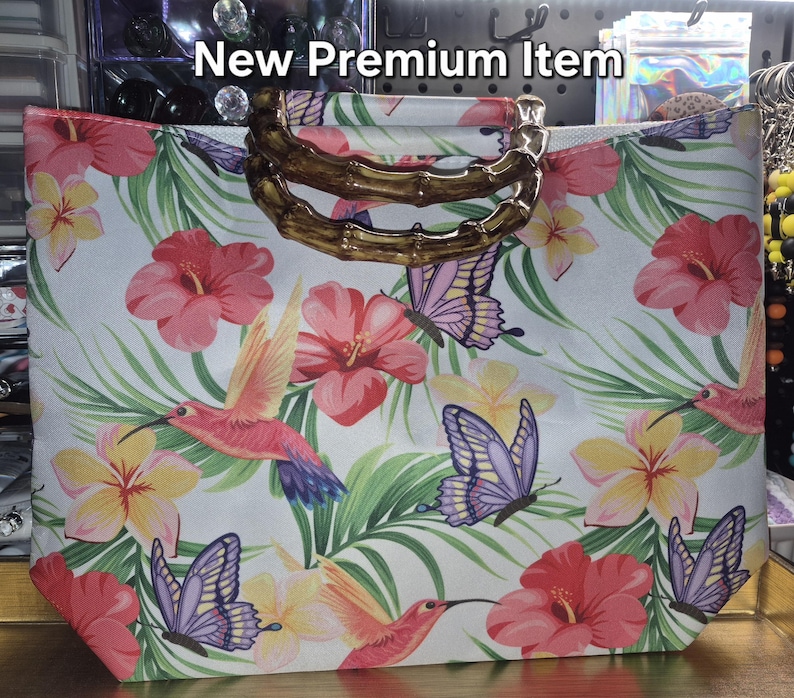 May include: A white tote bag featuring a vibrant tropical print of pink hibiscus flowers, yellow plumeria, green palm leaves, hummingbirds, and purple butterflies. The bag includes two curved, brown bamboo-style handles. Text reads New Premium Item.