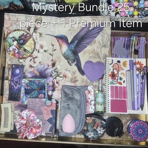 May include: A mystery bundle of 25 pieces plus 1 premium item displayed in a gold tray. Items include a floral hummingbird fabric, a stained glass hummingbird ornament, a purple heart note, a spiral notebook, pens, a scrunchie, and pouches.