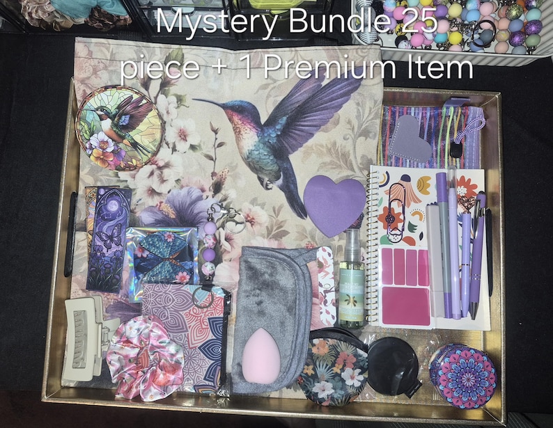 May include: A mystery bundle of 25 pieces plus 1 premium item displayed in a gold tray. Items include a floral hummingbird fabric, a stained glass hummingbird ornament, a purple heart note, a spiral notebook, pens, a scrunchie, and pouches.
