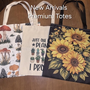 May include: Three tote bags with white handles. The left bag features a white background with various mushrooms. The centre bag is cream-coloured with a cactus graphic and the text "JUST ONE PLANT I PROMISE". The right bag is black with yellow sunflowers.