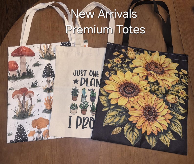 May include: Three tote bags with white handles. The left bag features a white background with various mushrooms. The centre bag is cream-coloured with a cactus graphic and the text "JUST ONE PLANT I PROMISE". The right bag is black with yellow sunflowers.