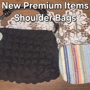 May include: Two shoulder bags with the text New Premium Items Shoulder Bags. One is a black textured bag with a light green flower-shaped tag and a black handle. The other is a rectangular bag with a multi-coloured striped pattern and thin straps.