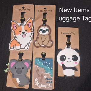 May include: Five luggage tags on brown cardboard backing labelled Luggage Tag. Designs include a corgi, a sloth, a koala on a branch, a beach scene with footprints and the text Peace Love Sandy Feel, and a panda. The text New Items Luggage Tags is visible.