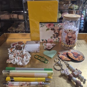 May include: A collection of stationery and accessories featuring a yellow notebook, a zippered pouch with a highland cow print, a glass tumbler with a bamboo lid, a scrunchie, two beaded keychains, five pens, stickers, a notepad, and coasters.