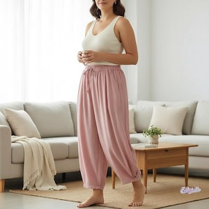 May include: A woman wearing a cream-colored tank top and loose, blush-pink pants with a drawstring waist. The pants are gathered at the ankles. The setting is a living room with a neutral-toned sofa and a wooden coffee table.