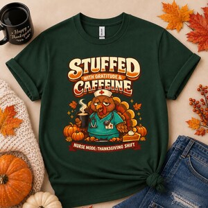 Op de afbeelding: Een donkergroen t-shirt met een afbeelding van een kalkoen verkleed als verpleegkundige met een stethoscoop en kapje, die een koffiekop vasthoudt. De tekst luidt STUFFED WITH GRATITUDE & CAFFEINE en NURSE MODE: THANKSGIVING SHIFT, omringd door pompoenen.