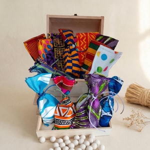 Könnte beinhalten: Eine Holzkiste und ein Tablett gefüllt mit bunten Stoffbeuteln, die mit Schnur gebunden sind. Die Beutel zeigen verschiedene Muster und Farben, darunter Orange, Blau, Lila und Weiß. Weiße Kugeln sind vor dem Tablett verstreut, mit einem Bündel Stroh und einem Zweig getrockneter Blumen im Hintergrund.