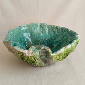 Peut inclure: Un bol en céramique fait à la main avec une forme organique unique. Le bol présente une surface texturée et est émaillé dans des tons de vert et de sarcelle, avec des touches de marron. Le bord a un design ondulé, ajoutant à son attrait artistique. Ce bol décoratif est adapté à la décoration intérieure.