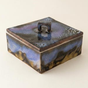 Op de afbeelding: Vierkante keramische doos met deksel en handvat. De doos heeft een glazuur in tinten blauw, paars en bruin. De handgreep is een klein, gebogen ontwerp. De doos is een decoratief opbergitem.